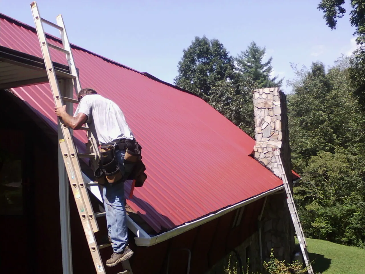Roofer on a ladder beside a red metal roof and stone chimney — Soffit & Fascia Repair by Roofing Brothers in Newport Coast